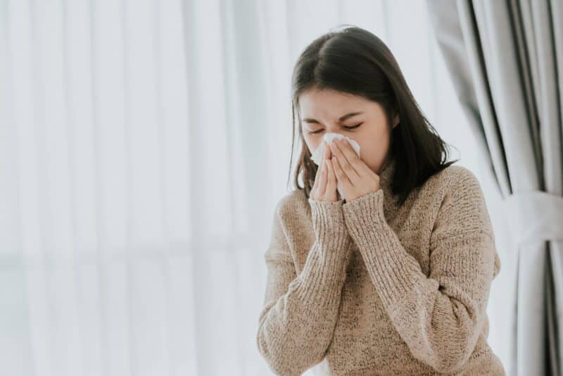 woman wearing a beige sweater blowing her nose into a white tissue